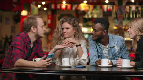 Fröhliche-Gesellschaft-Von-Freunden-In-Einer-Bar,-Die-Kaffee-Trinken,-Lachen-Und-Diskutieren-Und-Dabei-Auf-Den-Smartphone-Bildschirm-Schauen.